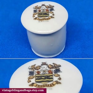 May include: White porcelain lidded jar with a gold and black crest featuring a bird and the word "Progress" on the lid.
