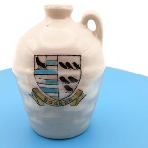 May include: A cream-colored ceramic jug with a single handle and a narrow neck. The front features a shield-shaped crest with blue and white horizontal stripes, black bird silhouettes, and a yellow banner underneath with the text BOGNOR.