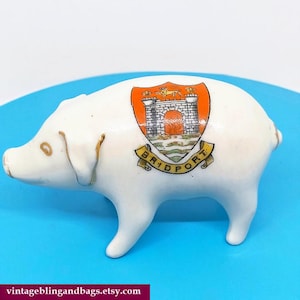 May include: White ceramic pig figurine with a colorful Bridport crest on its side. The crest features a castle, orange and yellow accents, and the town's name. The pig has gold-colored accents on its ears and snout.