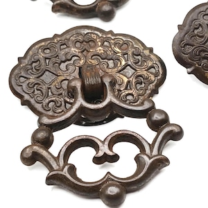 May include: A set of dark brown metal drawer pulls featuring an ornate, embossed scrollwork backplate and a matching drop-style handle. The hardware displays a weathered, aged finish with intricate, symmetrical patterns across the surface.