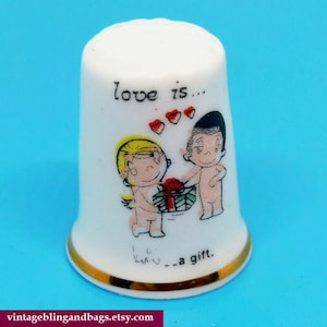 May include: White ceramic thimble with a gold rim. It features a cartoon couple with the text "love is... love... a gift."
