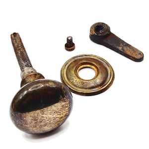 Könnte beinhalten: Ein antikes Türknauf-Set mit einem angelaufenen, oval geformten Knauf, einer runden Rosette, einer kleinen Schraube und einem Hebel. Die Metallkomponenten weisen ein abgenutztes, gealtertes Aussehen mit Braun- und Goldtönen auf, was auf Vintage-Hardware hindeutet.