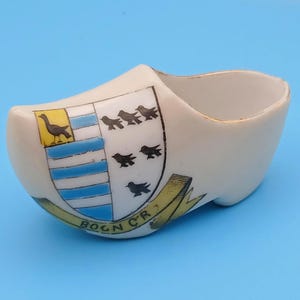 Puede incluir: Un pequeño zueco de cerámica blanca con un escudo decorativo que presenta un cuadrado amarillo con un pájaro, rayas azules y blancas y pájaros negros. El zueco tiene un ribete dorado y el texto "BOGN CR" en una pancarta amarilla.