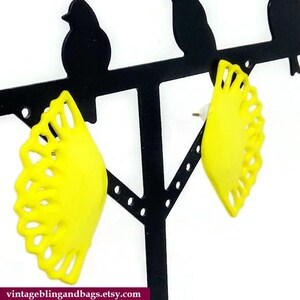May include: A pair of yellow, fan-shaped earrings with a delicate, lacy design. The earrings are displayed on a black earring stand.