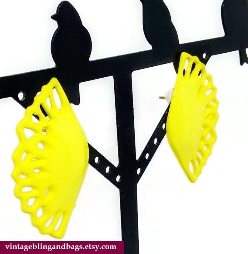 May include: A pair of yellow, fan-shaped earrings with a delicate, lacy design. The earrings are displayed on a black earring stand.