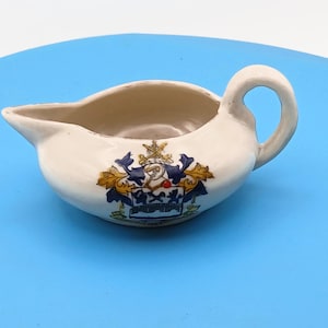 May include: A small, cream-colored ceramic gravy boat featuring a handle on the right side and a pouring spout on the left. The front of the vessel is decorated with a colorful heraldic crest displaying blue, gold, and white elements.