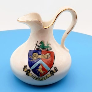 May include: A small white ceramic creamer pitcher featuring a gold-trimmed rim and handle. The front displays a colorful heraldic crest with a lion, a castle, and a horse, with a banner below the shield bearing the text MARGATE in black letters.