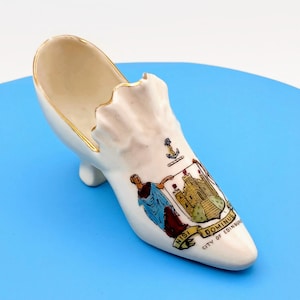 May include: A cream-colored porcelain shoe figurine featuring a gold-trimmed ruffled edge. The front displays a colorful crest with a castle, an anchor, and a figure in a blue robe, with the text NISI DOMINUS and CITY OF EDINBURGH printed below.