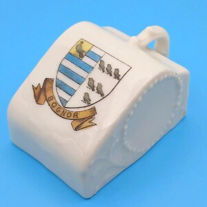 Puede incluir: Cenicero de cerámica blanca con asa y diseño decorativo. El cenicero presenta un escudo con rayas azules y blancas, pájaros y la palabra "BOGNOR" en una pancarta. El cenicero tiene un borde redondeado con un patrón de puntos.