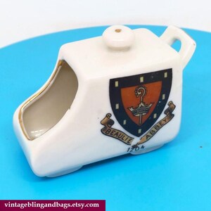 May include: White ceramic sugar bowl shaped like a small car with a gold trim. The bowl has a crest with the words "Beaulie Abbey 1204" on the side.