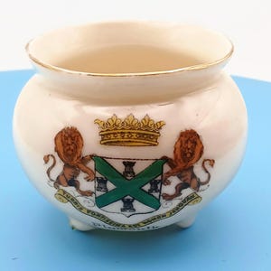 May include: A small, cream-colored ceramic pot with a gold-trimmed rim and three short feet. The front features a heraldic crest with two brown lions, a gold crown, and a green saltire cross. Below, a banner reads: TURRIS FORTISSIMA EST NOMEN JEHOVAH.