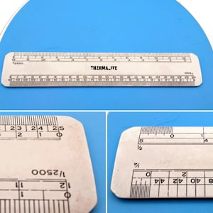 May include: A silver-colored metal architectural scale ruler featuring the brand name THERMAJITE printed in black. The ruler displays multiple measurement scales, including 1/2500 and 1/500, with clear numerical markings and graduation lines.