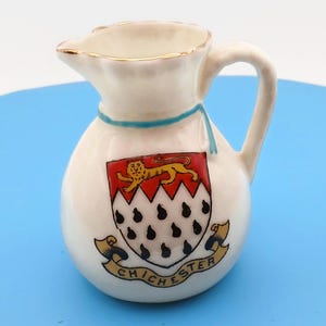 May include: A small white ceramic pitcher with a gold-trimmed rim and a handle. The body features a painted coat of arms with a gold lion on a red field above a white field with black ermine spots. A blue ribbon is painted around the neck, and the word CHICHESTER is printed on a gold banner below the shield.