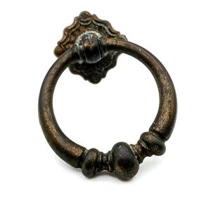Puede incluir: Tirador de cajón en tono bronce antiguo con placa trasera decorativa y asa de anillo. El herraje presenta un acabado envejecido, añadiendo una estética vintage. Ideal para armarios y cómodas.
