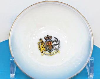 May include: White ceramic saucer with a gold rim and a colorful royal crest in the center. The crest features a lion and a unicorn flanking a shield with the text "Coronation 1953".