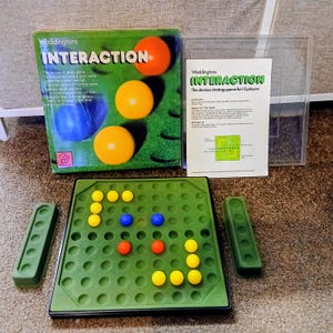 May include: A vintage board game called "Interaction" by Waddingtons. The box features the game's title and colorful spheres. The game includes a green playing board with holes, colored balls, and two green game pieces. The game is for 1-2 players.