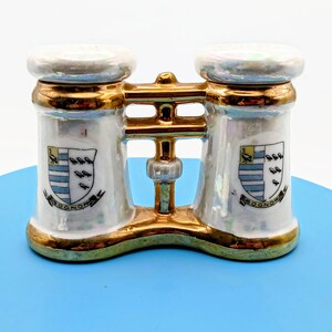 May include: A vintage, white porcelain salt and pepper shaker set shaped like binoculars. The set features gold trim, a pearlescent finish, and a crest with the word "Bognor". The crest includes a bird, stripes, and other small symbols.