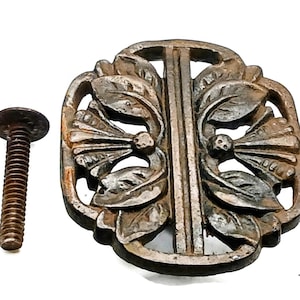 Könnte beinhalten: Ein verwitterter, dunkelmetallischer Schubladengriff mit einem symmetrischen, durchbrochenen Design aus stilisierten Blättern und zentralen vertikalen Stäben, begleitet von einer passenden Gewindeschraube mit flachem Kopf.