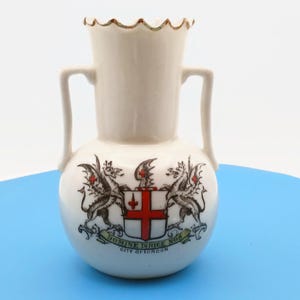 Puede incluir: Un pequeño jarrón de cerámica blanquecina con dos asas. Presenta un escudo con dos dragones, una cruz roja y el texto "DOMINE DIRIGE NOS CITY OF LONDON". El borde tiene un diseño festoneado.