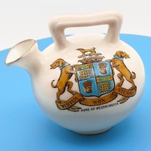 May include: Cream-colored ceramic pitcher with a handle and spout, featuring a coat of arms with two golden dogs, a shield, and the text "VIRTUS NON STEMMA DUKE OF WESTMINSTER". The spout has a gold rim.