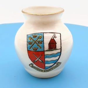 Puede incluir: Un pequeño jarrón de cerámica de color crema con un borde dorado. La parte frontal presenta un escudo heráldico colorido dividido en cuatro cuadrantes, que muestra coronas, bastones cruzados, un edificio rojo y ondas azules, con el texto BISHOPS STORTFORD.