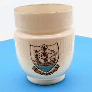 May include: Cream-colored ceramic cup with a gold rim and a decorative crest featuring a ship, crown, and lion. The cup has a ridged texture and the text "F.LDEBURGH" on a banner below the crest.