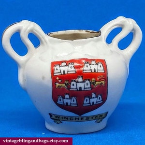 Peut inclure: Un vase en céramique blanche avec deux anses en forme de cou de cygne. Le vase présente un blason rouge et or avec le mot "Winchester" imprimé en dessous.