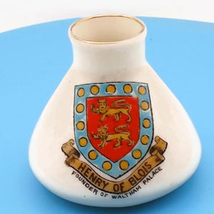 May include: A small white ceramic vase with a gold-trimmed rim. The front features a crest with two yellow lions on a red background, surrounded by a blue border with yellow circles. Below the crest, a banner reads HENRY OF BLOIS FOUNDER OF WALTHAM PALACE.
