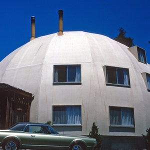 May include: A unique, dome-shaped house with multiple windows and two chimneys. A vintage green car is parked in front of the house. The house is painted a light beige color and has a wooden porch.