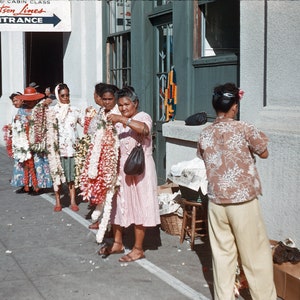 May include: Five women wearing floral leis stand in front of a building. They are all smiling and holding leis in their hands. The building has a sign that says "Matson Lines" and "First & Cabin Class Entrance".