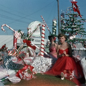 May include: A festive Christmas parade float decorated with candy canes, red bows, and a small Christmas tree. A woman in a red dress sits on the float, smiling.