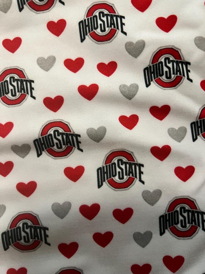 May include: A white fabric featuring a repeating pattern of the Ohio State University logo in red, black, and white, interspersed with numerous small solid red hearts and smaller solid grey hearts scattered across the entire surface.
