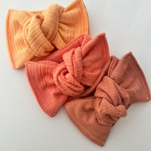 May include: Three knotted headbands in shades of orange and peach. The headbands are made of a ribbed fabric with a textured appearance. The headbands are arranged on a white surface. The headbands are a stylish accessory.