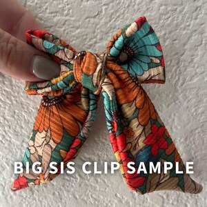 May include: A floral patterned hair bow with orange, teal, red, and yellow flowers. The text "BIG SIS CLIP SAMPLE" is printed in white. The bow is made of fabric and is tied in a bow shape.