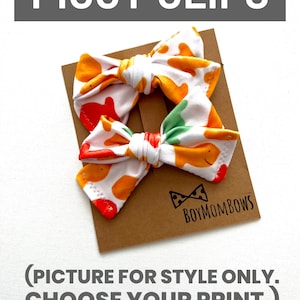 May include: Two fabric hair bows with a white background and a pattern of orange and yellow shapes. The bows are attached to a brown card with the text "PIGGY CLIPS" at the top and "BOYMOMBOWS" at the bottom. The text "(PICTURE FOR STYLE ONLY. CHOOSE YOUR PRINT.)" is also visible.