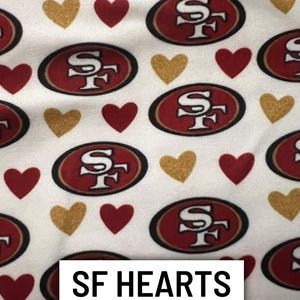 Może przedstawiać: Biała tkanina z powtarzającym się wzorem czerwonych i złotych serc oraz czerwonych i złotych logo SF. Tekst "SF HEARTS" jest wyświetlany w białym prostokącie.