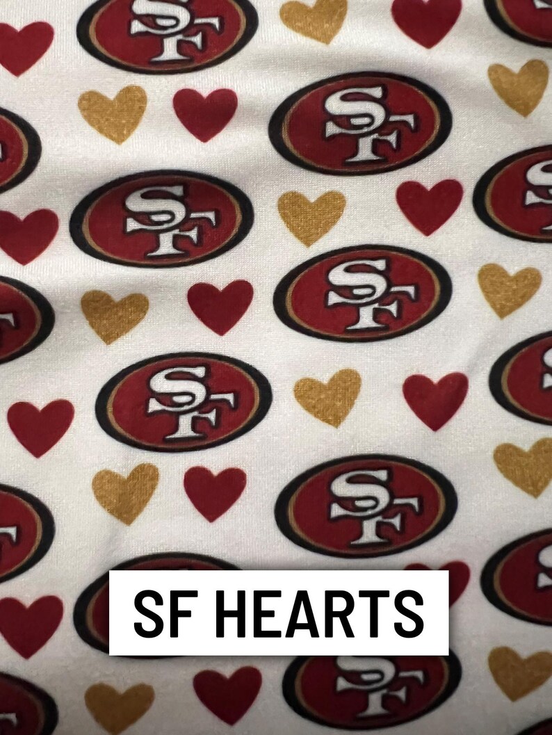 Może przedstawiać: Biała tkanina z powtarzającym się wzorem czerwonych i złotych serc oraz czerwonych i złotych logo SF. Tekst "SF HEARTS" jest wyświetlany w białym prostokącie.