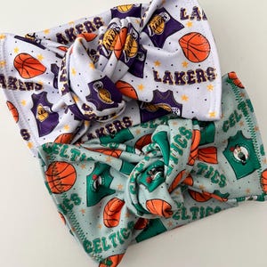 May include: Two knotted headbands featuring basketball team logos. One headband is white with purple and gold Los Angeles Lakers logos and basketballs. The other headband is light blue with green and orange Boston Celtics logos and basketballs.