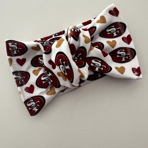 Może przedstawiać: Biała, wiązana opaska na głowę z logo San Francisco 49ers w kolorze czerwonym i złotym, wraz z czerwonymi i złotymi kształtami serc. Opaska wykonana jest z miękkiego, elastycznego materiału.