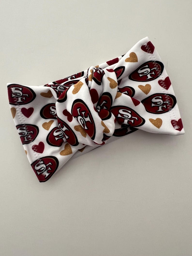 Może przedstawiać: Biała, wiązana opaska na głowę z logo San Francisco 49ers w kolorze czerwonym i złotym, wraz z czerwonymi i złotymi kształtami serc. Opaska wykonana jest z miękkiego, elastycznego materiału.
