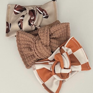 May include: Three knotted headbands in a neutral color palette. One headband features a football print, another is a solid brown knit, and the third has a checkered pattern in orange and white. The headbands are made of soft, ribbed fabric.