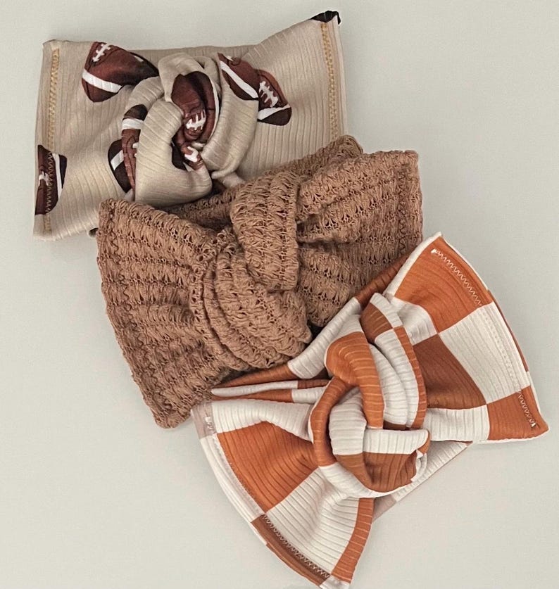 May include: Three knotted headbands in a neutral color palette. One headband features a football print, another is a solid brown knit, and the third has a checkered pattern in orange and white. The headbands are made of soft, ribbed fabric.