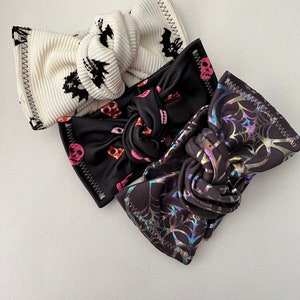 May include: Three Halloween-themed headbands. One is white with black bats, one is black with pink and orange skulls, and one is black with a holographic spiderweb design.