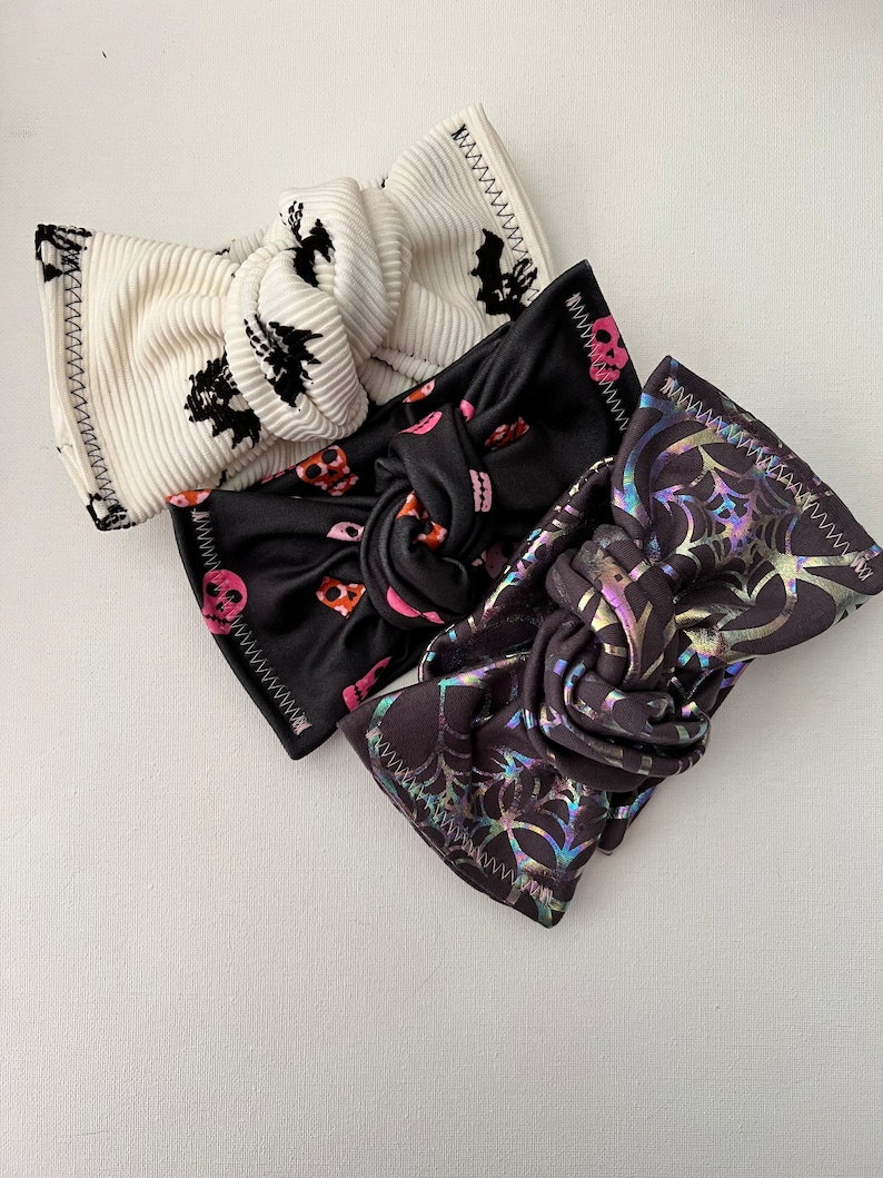 May include: Three Halloween-themed headbands. One is white with black bats, one is black with pink and orange skulls, and one is black with a holographic spiderweb design.