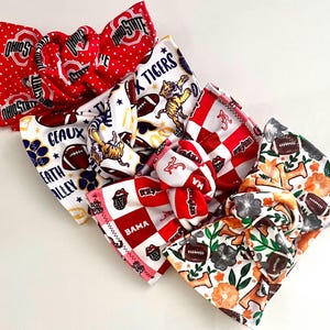 May include: A collection of four fabric headbands featuring various sports team themes. One is red with white polka dots and Ohio State logos. Another is white with tiger graphics and footballs. A third is red and white checkered with Bama text. The last is white with orange flowers and footballs.