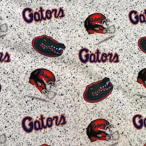 May include: A white fabric with a black speckled pattern featuring a repeating print of the word Gators in blue, orange football helmets with the word Gator, and green alligator head logos with red and blue outlines throughout the material.