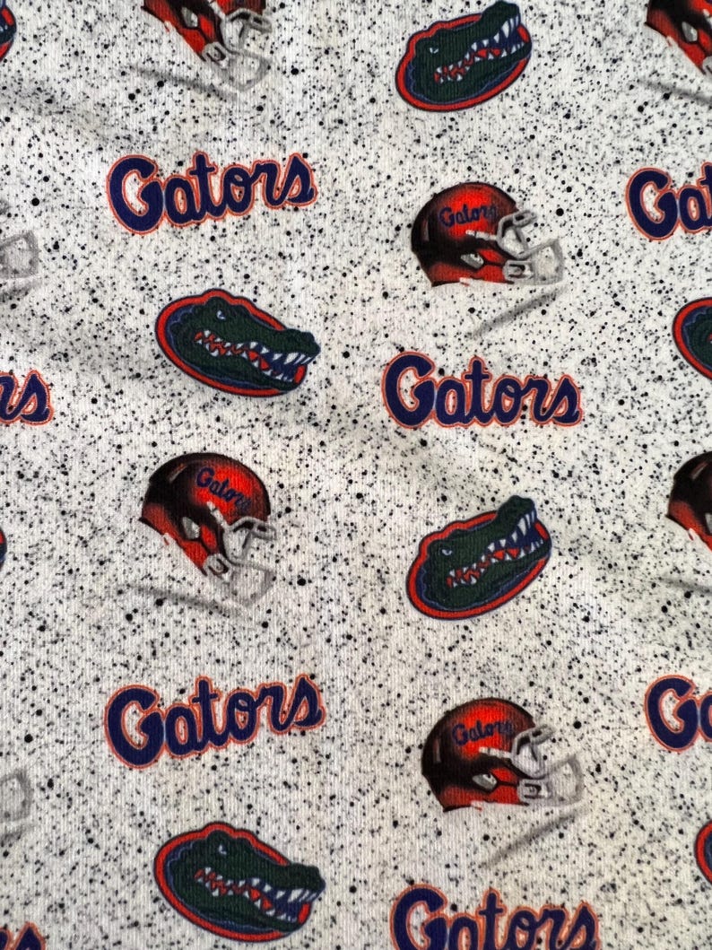 May include: A white fabric with a black speckled pattern featuring a repeating print of the word Gators in blue, orange football helmets with the word Gator, and green alligator head logos with red and blue outlines throughout the material.