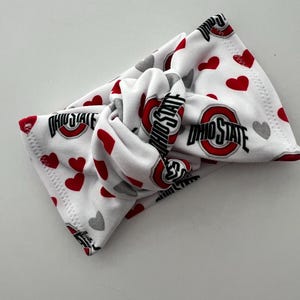 May include: A white fabric headband featuring a knotted center design. The pattern consists of repeating red hearts, grey hearts, and the Ohio State logo with the text OHIO STATE in black and red lettering printed across the soft material surface.