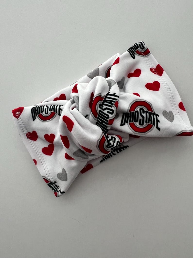 May include: A white fabric headband featuring a knotted center design. The pattern consists of repeating red hearts, grey hearts, and the Ohio State logo with the text OHIO STATE in black and red lettering printed across the soft material surface.