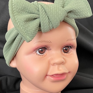 May include: Sage green baby headband with a large bow. The headband is made of a textured fabric and is displayed on a mannequin head with brown eyes and a smiling expression. The bow is centered on the head.
